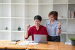 © PHAISITSAWAN - Cheerful business people using a laptop in an office. Happy young entrepreneurs smiling while working together in a modern workspace. Two young business people are sitting together at a table.