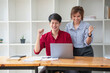 © PHAISITSAWAN - Cheerful business people using a laptop in an office. Happy young entrepreneurs smiling while working together in a modern workspace. Two young business people are sitting together at a table.