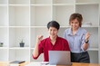 © PHAISITSAWAN - Cheerful business people using a laptop in an office. Happy young entrepreneurs smiling while working together in a modern workspace. Two young business people are sitting together at a table.