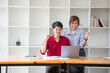 © PHAISITSAWAN - Cheerful business people using a laptop in an office. Happy young entrepreneurs smiling while working together in a modern workspace. Two young business people are sitting together at a table.