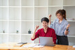 © PHAISITSAWAN - Cheerful business people using a laptop in an office. Happy young entrepreneurs smiling while working together in a modern workspace. Two young business people are sitting together at a table.
