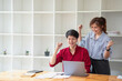 © PHAISITSAWAN - Cheerful business people using a laptop in an office. Happy young entrepreneurs smiling while working together in a modern workspace. Two young business people are sitting together at a table.