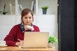 © PHAISITSAWAN - Beautiful Asian businesswoman sitting at a table with computer wearing headphones with microphone Looking at the laptop camera Happy businesswoman educating customers online.
