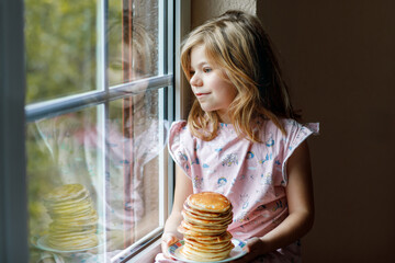 Wall Mural - Little happy preschool girl with a large stack of pancakes for breakfast. Positive child eating healthy homemade food in the morning.