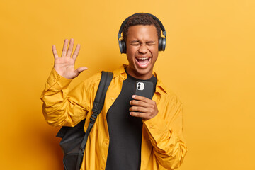 Sticker - Overjoyed dark skinned man holds smartphone listens music via headphones and exclaims loudly dressed in casual clothing carries rucksack on shoulder isolated over yellow background. Hobby concept