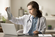 Serious Young Practitioner Woman Looking At Xray Scan Image Of Bones, Sitting At Workplace Table With Laptop In Doctor Office, Studying Radiography Examination Result, Thinking On Diagnosis