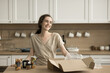 © fizkes - Happy pretty adult girl unpacking cardboard box with canned vegetables in kitchen, getting grocery purchase from supermarket, standing at table with glass jars, smiling, laughing
