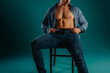 Fit shirtless guy with unbuttoned shirt sitting on a chair posing in studio on a turquoise background