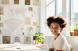 © Kate Simon - A 10-year-old afro-american girl sits at a desk in a white sunny classroom in a European learning center, on the white wall of which children's drawings hang