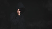 An Unknown Man In Hood Jacket Using Cellphone In Dark Background. Hacker, Scammer Or Criminal Using Smartphone To Lure Victim, Cyber Criminal Concept.
