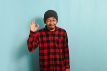 Smiling, Winking Young Asian Man Does The OK Sign Gesture Says Good Job, Isolated On Blue Background