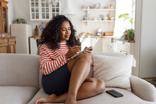 Cute barefoot african american woman of 20s sitting on cozy sofa at home against kitchen background, enjoying leisure time, writing in her diary, noting down ideas or planning next weekend