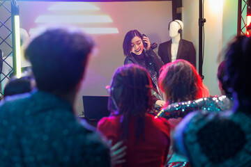 Crowd watching musician performance while attending discotheque party in nightclub. Young asian woman dj playing using headphones and digital controller on stage with spotlights in club