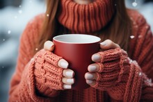 a red mug in the hands of those dressed in knitted mittens against the backdrop of a blurred snow landscape