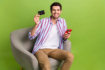 Sticker - Portrait of toothy beaming person wear stylish clothes sit in armchair hold smartphone debit card isolated on green color background