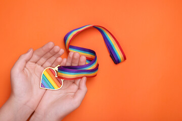 Woman holding rainbow ribbon with heart shaped pendant on orange background, top view and space for text. LGBT pride
