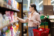 © _KUBE_ - Sale in grocery store. Side view of young caucasian woman wearing eyeglasses holds cart and take food from upper shelf. Concept of shopping in supermarket and consumerism