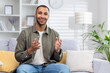 © Tetiana - Portrait of a young African-American man making a video call from home, sitting on a sofa in front of the camera and smilingly gesturing with his hands