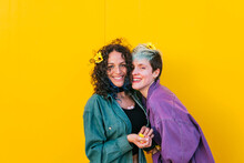 Happy Lesbian Couple With Flowers On Hair Standing In Front Of Yellow Wall