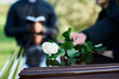 © pressmaster - Fresh white rose being put by woman in mourning attire on top of closed coffin lid against priest with open Bible carrying out funeral service