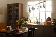 Bunch Of Yellow Wildflowers In Vase, Ripe Vegetables And Other Stuff On Wooden Table Standing In The Center Of Spacious Kitchen Of Cottage