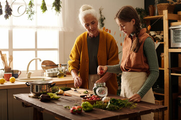 Cute girl in apron putting fresh tomato into open jar with marinade mix while helping her grandmother prepare vegetables for winter