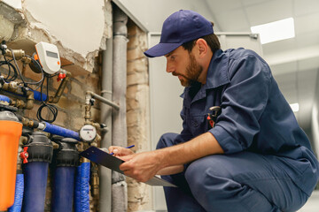 Male plumber diagnoses pipes in water supply system before repairing and makes notes in clipboard