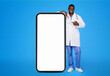 © Prostock-studio - Smiling young black guy doctor in white coat point finger at big smartphone with empty screen