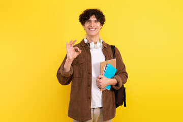 Student man with notebooks and backpack gestures okay, yellow background