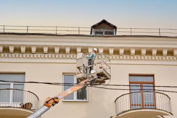 Wall Mural - Man paint wall with spray gun on aerial lifting platform. Painter working in crane bucket, paint building wall of historic building with paint spray gun. Repair and restoration facade of building.