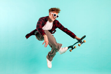 Wall Mural - young crazy guy rides skateboard and jumps on blue isolated background, hipster in sunglasses flies