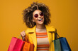 © Sascha - Shopping Lifestyle: Joyful young shopper posing with her shopping bags, expressing happiness against a yellow-orange studio background.