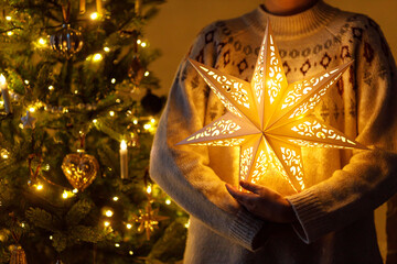 Wall Mural - Merry Christmas! Stylish illuminated golden star in hands on background of modern decorated christmas tree with golden lights in evening room. Atmospheric magical christmas eve