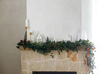 Wall Mural - Stylish decorated christmas fireplace. Modern christmas trees, houses, pine cones and spruce branches on fireplace mantel with brass bells and ribbon. Christmas scandinavian living room decor