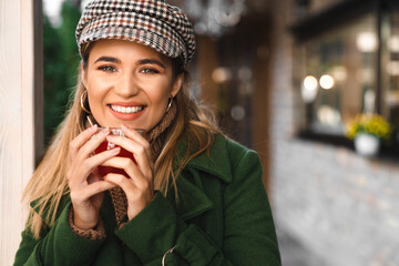 Wall Mural - Close up of blonde happy young woman looking at camera while holding cup of tea. Portrait of young beautiful smiling woman holding cup of tea with blurred background.