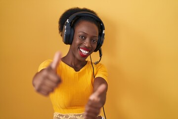 Wall Mural - African woman with curly hair standing over yellow background wearing headphones approving doing positive gesture with hand, thumbs up smiling and happy for success. winner gesture.