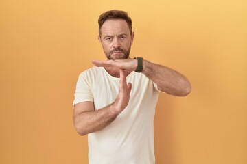 Poster - Middle age man with beard standing over yellow background doing time out gesture with hands, frustrated and serious face