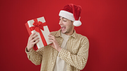 Wall Mural - Young hispanic man smiling confident wearing christmas hat holding gift over isolated red background