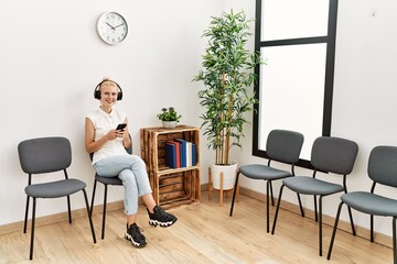 Poster - Young blonde woman using smartphone and headphones sitting on chair at waiting room