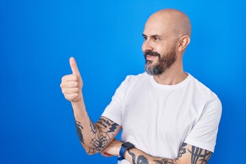 Sticker - Hispanic man with tattoos standing over blue background looking proud, smiling doing thumbs up gesture to the side