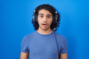 Canvas Print - Hispanic man with curly hair listening to music using headphones afraid and shocked with surprise expression, fear and excited face.