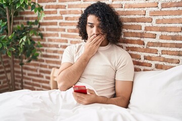 Wall Mural - Hispanic man with curly hair using smartphone sitting on the bed covering mouth with hand, shocked and afraid for mistake. surprised expression