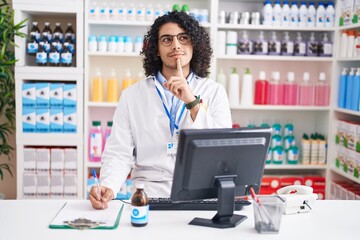 Poster - Hispanic man with curly hair working at pharmacy drugstore thinking concentrated about doubt with finger on chin and looking up wondering