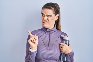 Sticker - Beautiful woman wearing sportswear holding water bottle pointing aside worried and nervous with forefinger, concerned and surprised expression