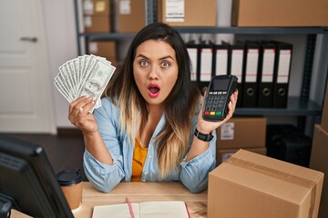 Poster - Young hispanic woman working at small business ecommerce holding money and dataphone afraid and shocked with surprise and amazed expression, fear and excited face.