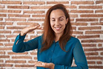 Canvas Print - Brunette woman standing over bricks wall gesturing with hands showing big and large size sign, measure symbol. smiling looking at the camera. measuring concept.