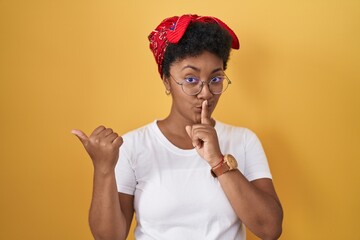 Poster - Young african american woman standing over yellow background asking to be quiet with finger on lips pointing with hand to the side. silence and secret concept.