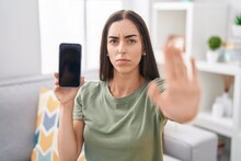 Young Brunette Woman Holding Smartphone Showing Blank Screen With Open Hand Doing Stop Sign With Serious And Confident Expression, Defense Gesture