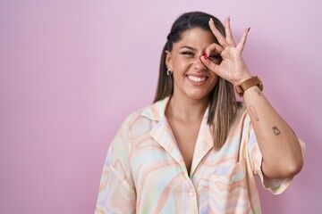 Wall Mural - Blonde woman standing over pink background doing ok gesture with hand smiling, eye looking through fingers with happy face.