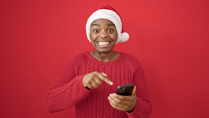 Wall Mural - African american woman wearing christmas hat pointing to smartphone over isolated red background